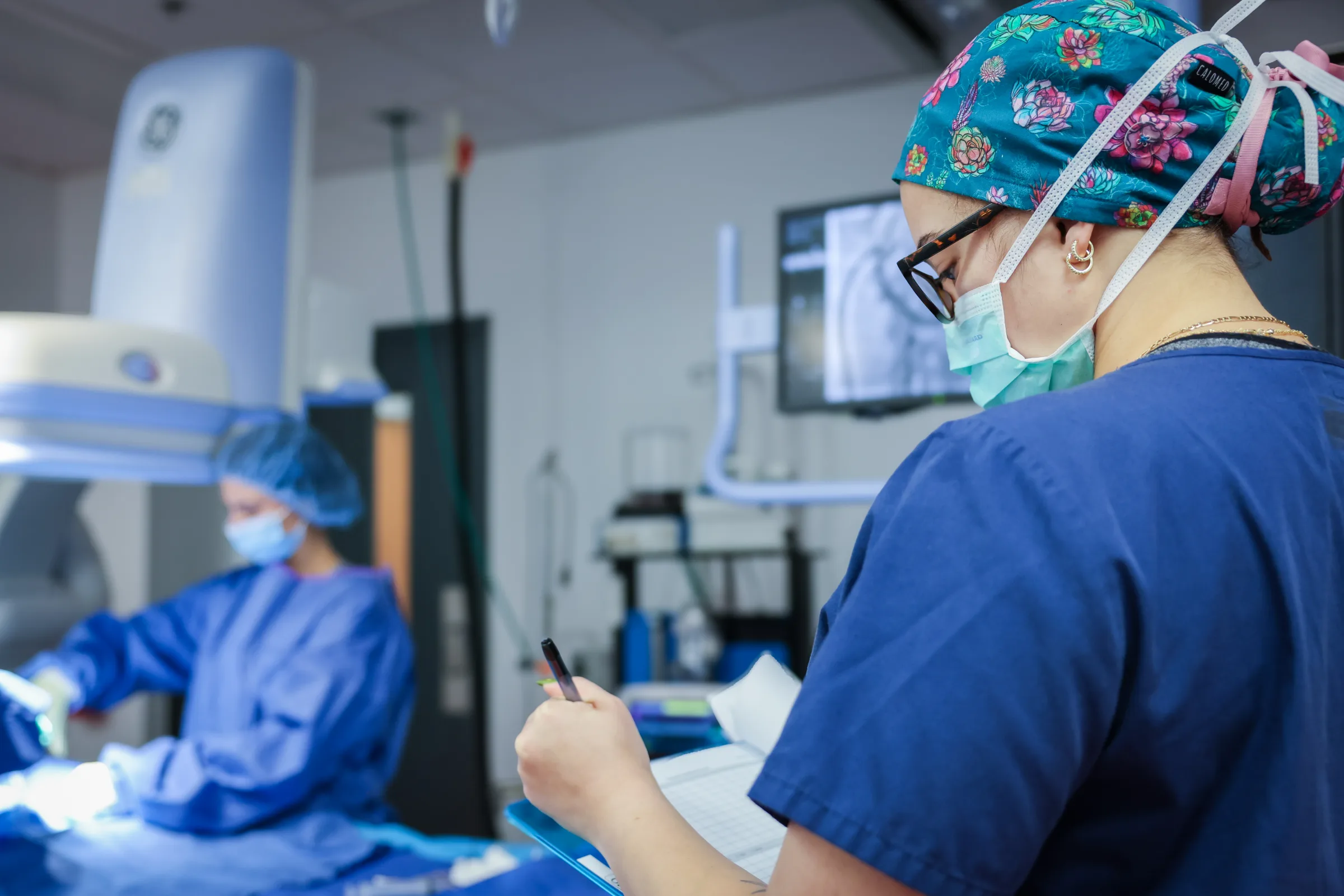 A Synchrony Labs team member recording study notes while a procedure continues behind her