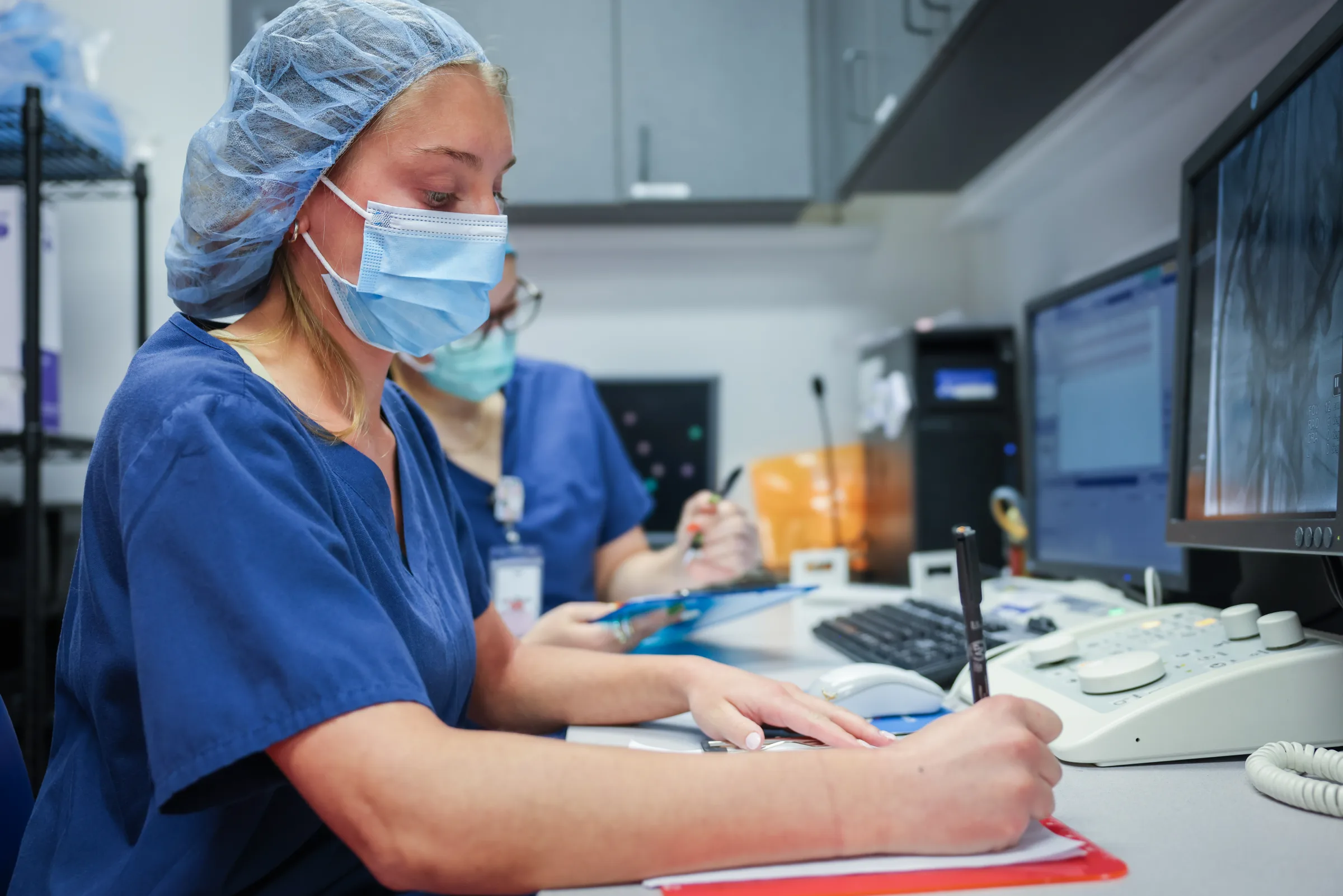 Synchrony Labs staff documenting a study at the control desk beside angiography monitors